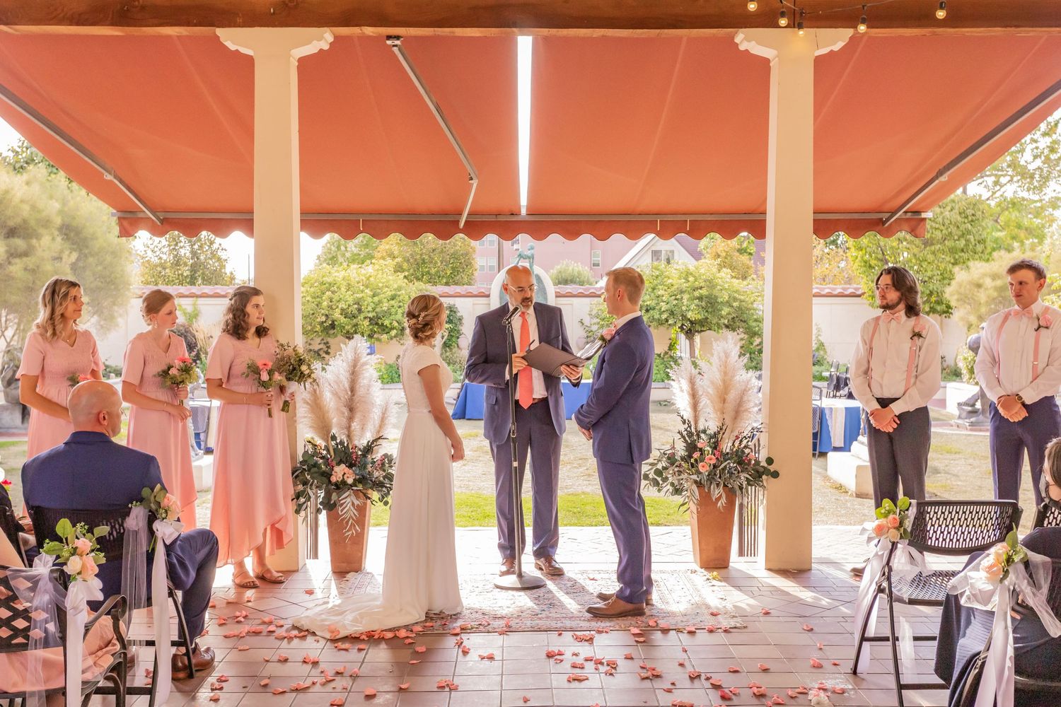 Wedding ceremony taking place under a covered outdoor patio with pink bridesmaid dresses and formal attire.