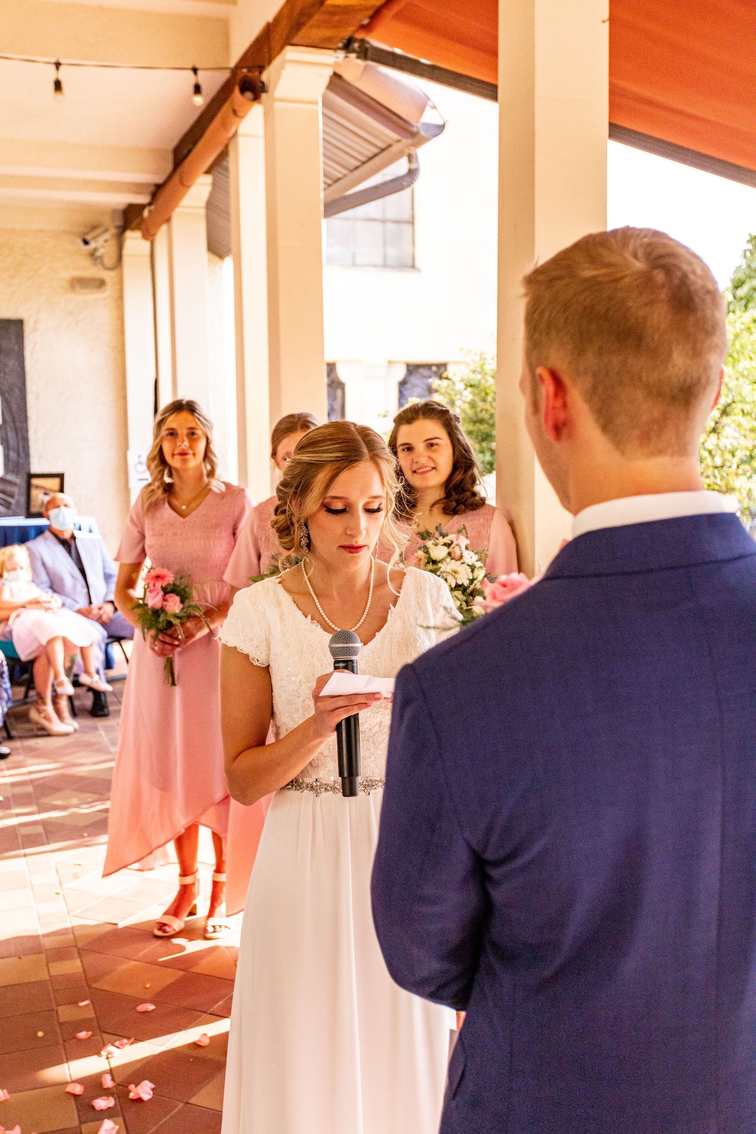 Wedding couple exchanging vows under a covered patio with bridesmaids in pink dresses looking on.