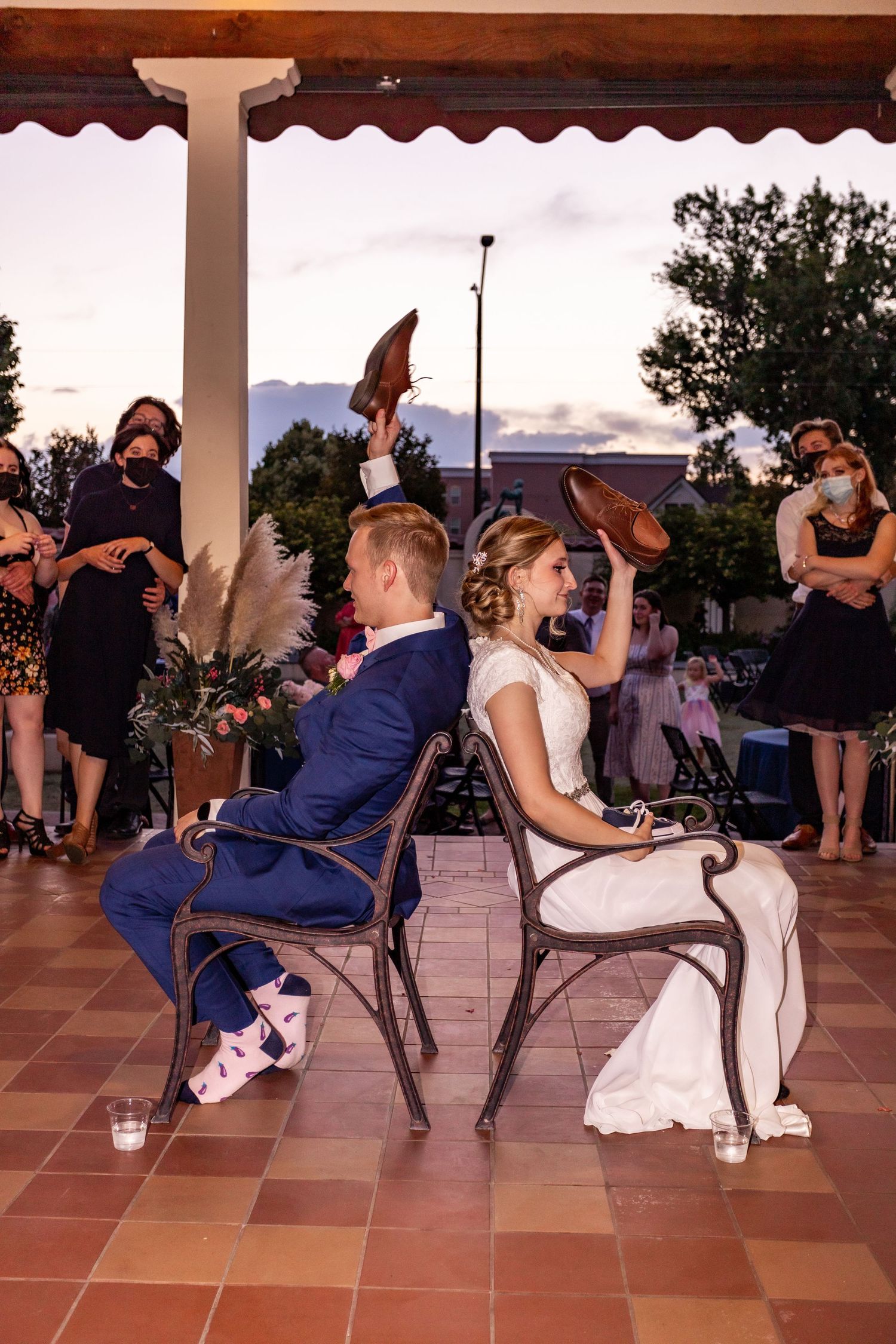 Wedding couple plays reception game sitting back-to-back on chairs under covered patio.