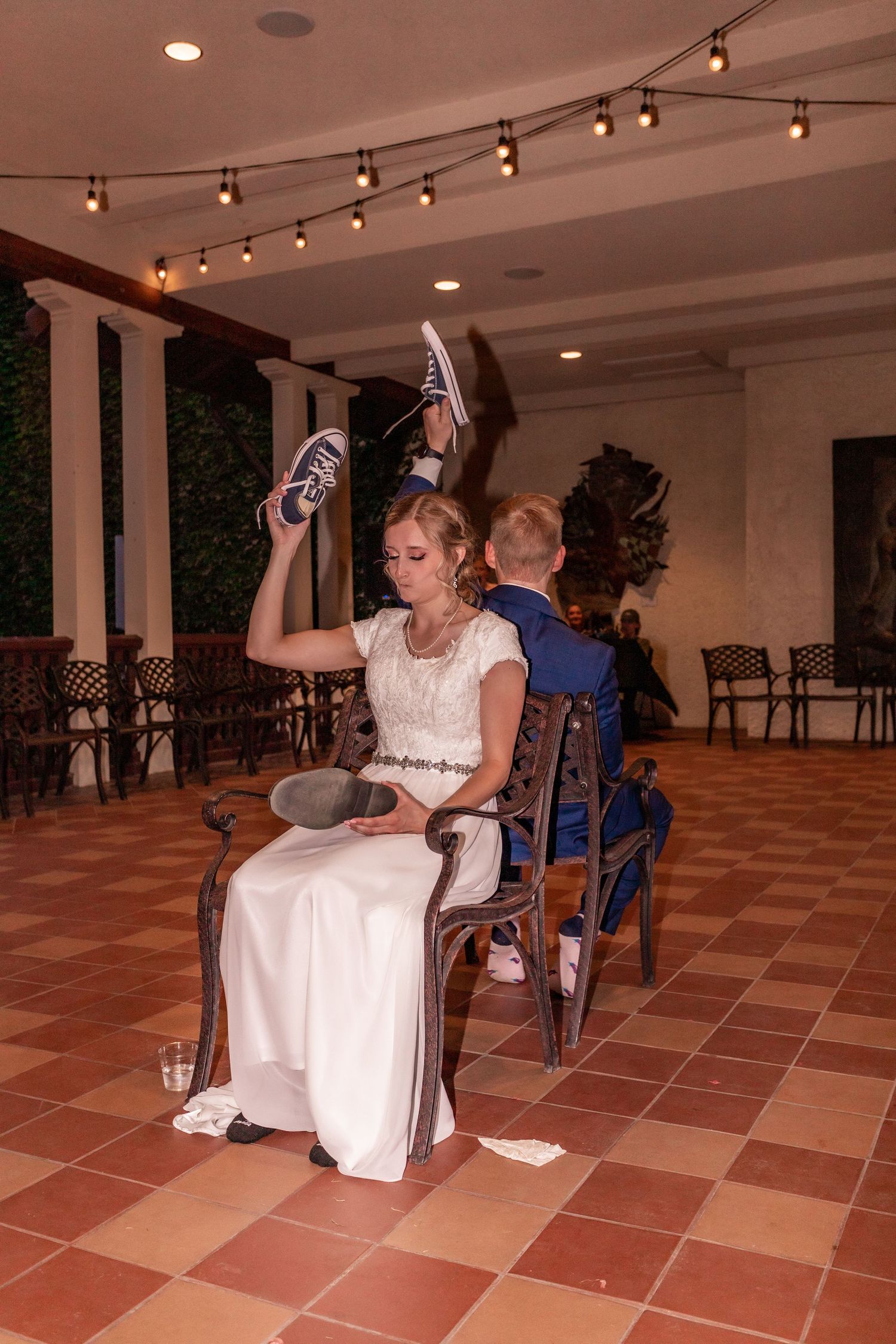 Wedding reception game where bride holds up shoes while seated on a chair under string lights.