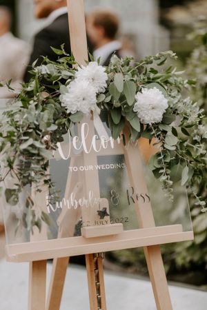 Wedding welcome sign with white flowers and greenery on wooden easel.