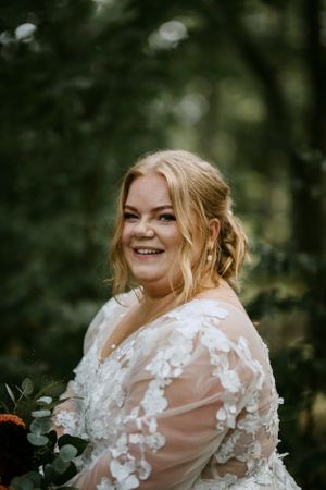 Bride wearing lace sleeve wedding dress smiles in woodland setting.