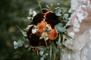 Wedding bouquet with deep burgundy and orange flowers against lace dress.