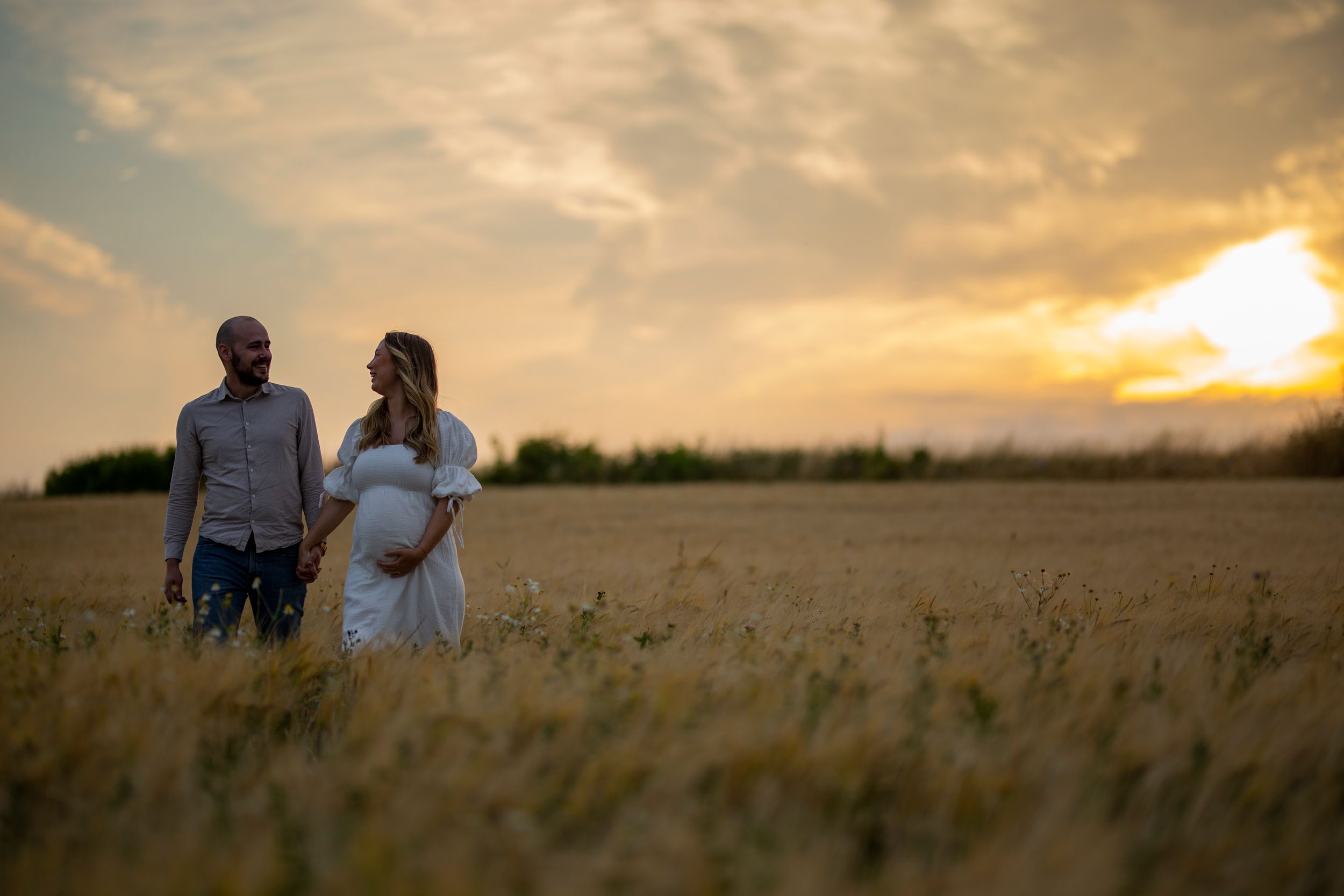 A couple stands together in a wheat field silhouetted against a golden sunset.