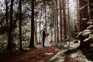 A romantic wedding photo of a couple standing on a forest path surrounded by tall evergreen trees.