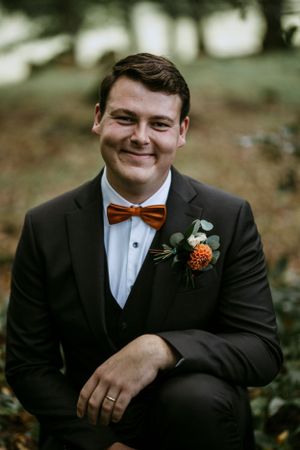 Groom in dark suit with brown bow tie and orange boutonniere smiles.