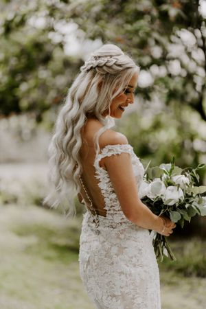 Bride in lace dress with braided hairstyle holds white bouquet in garden setting.