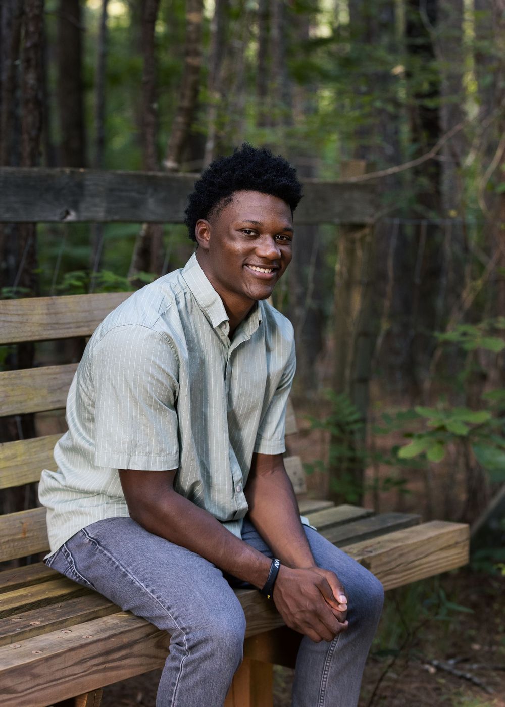 A relaxed outdoor portrait on a wooden bench surrounded by forest scenery.