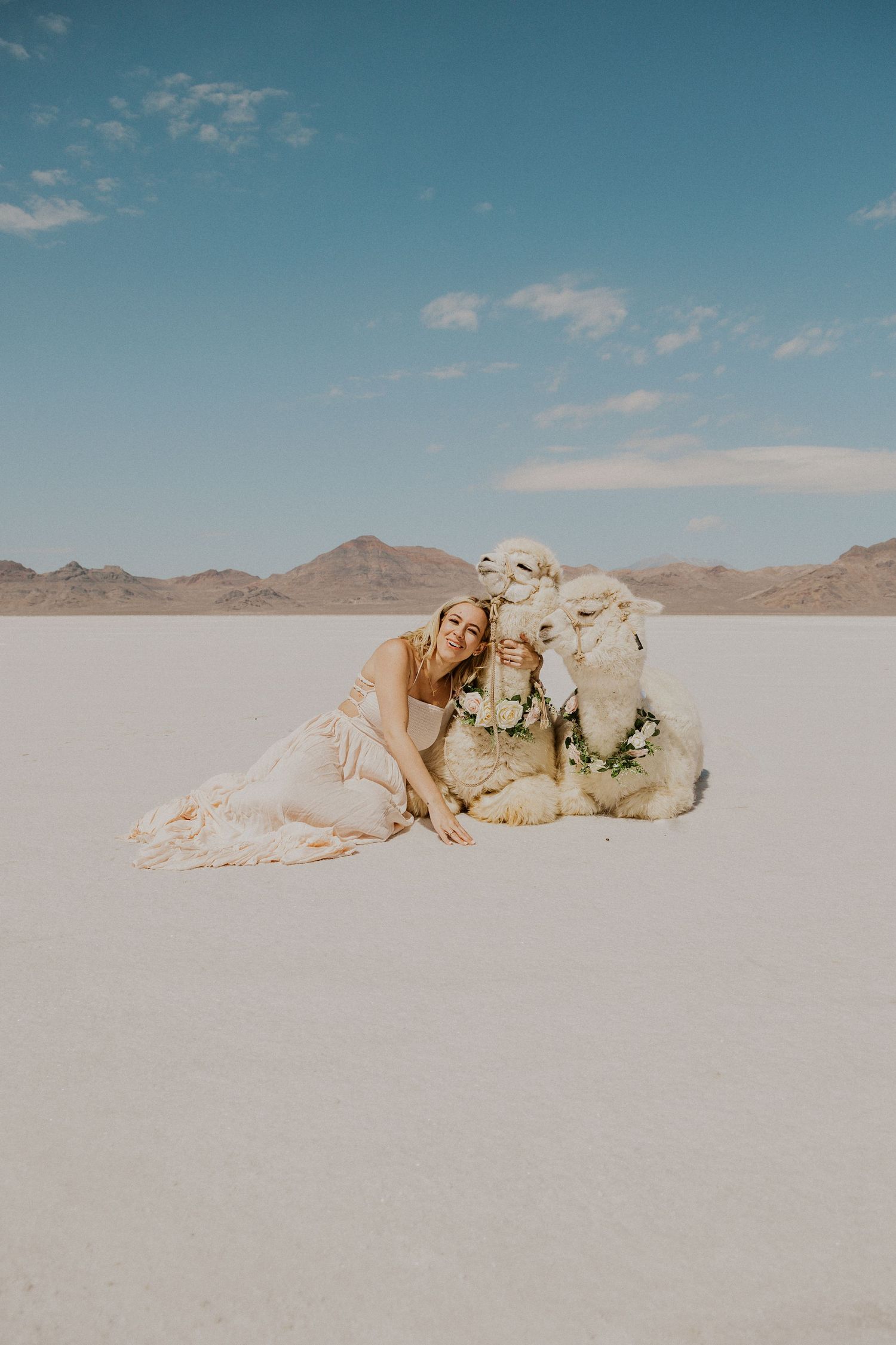 Woman in pink maxi dress snuggling up to two white alpacas at the bonneville salt flats. Alpacas are wearing floral necklaces.
