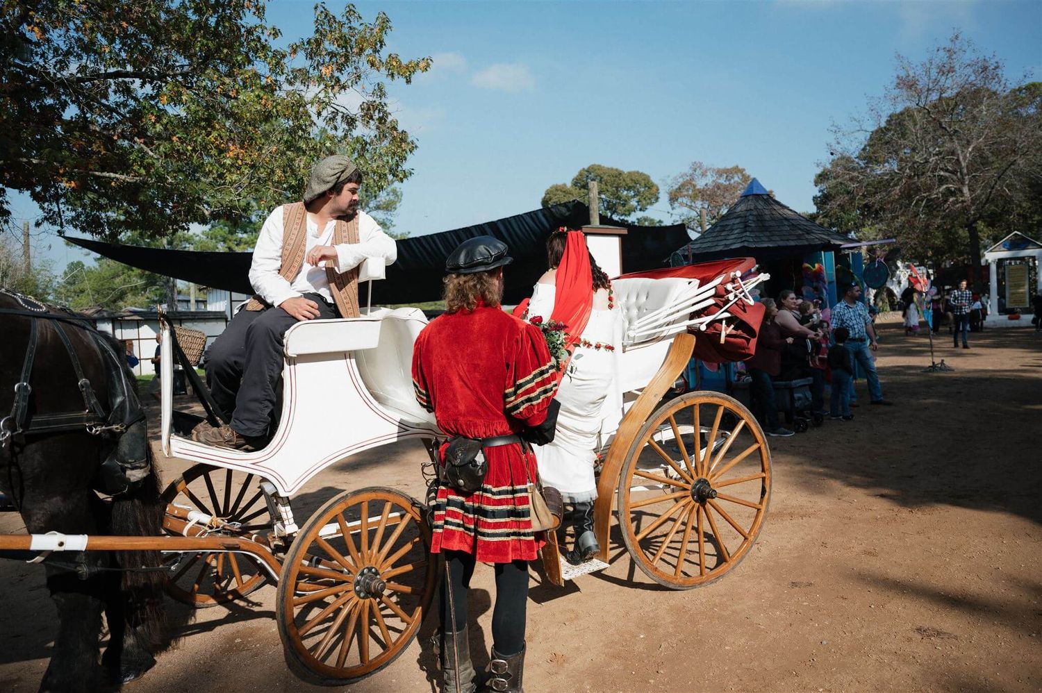 A white horse-drawn carriage with period-dressed attendants stands in a dusty renaissance faire setting.