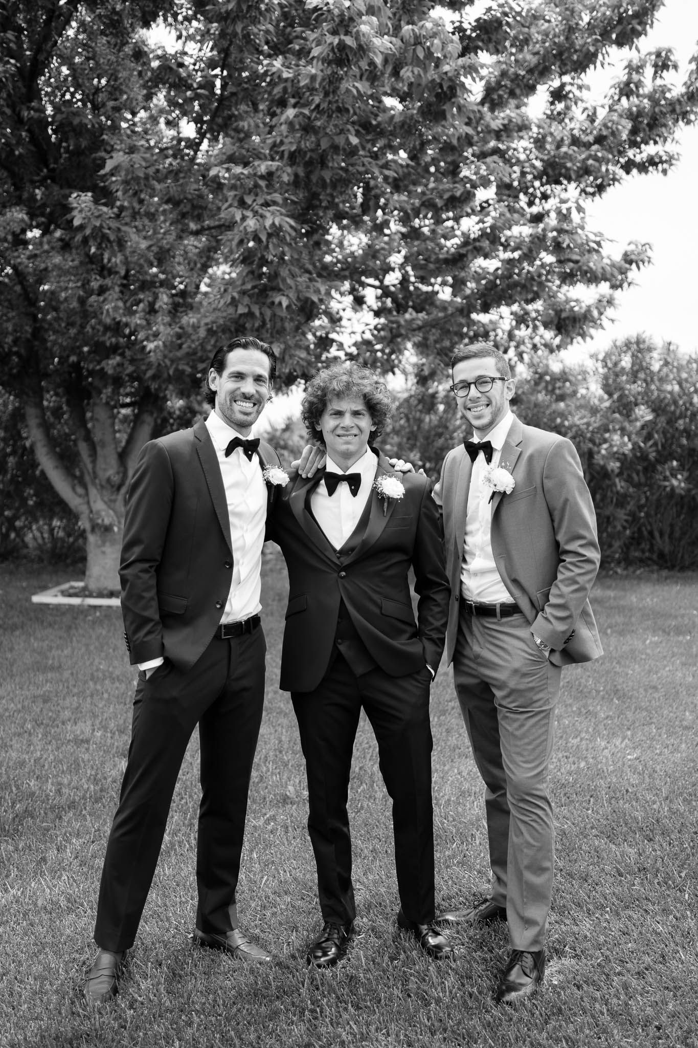 Three groomsmen in formal tuxedos pose together outdoors for a black and white wedding photo.