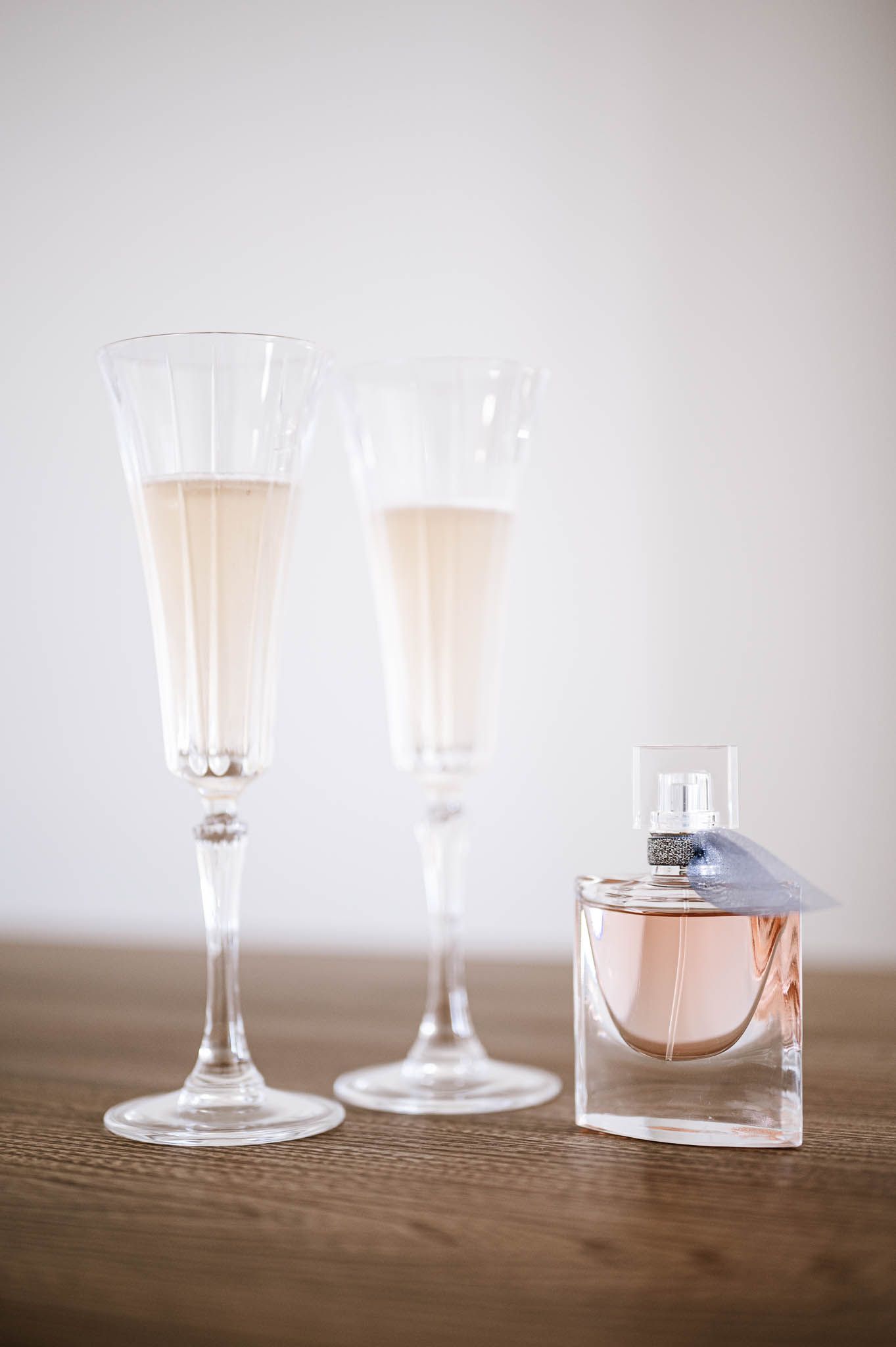 Two champagne flutes filled with blush-colored drinks and a perfume bottle arranged on wooden surface.