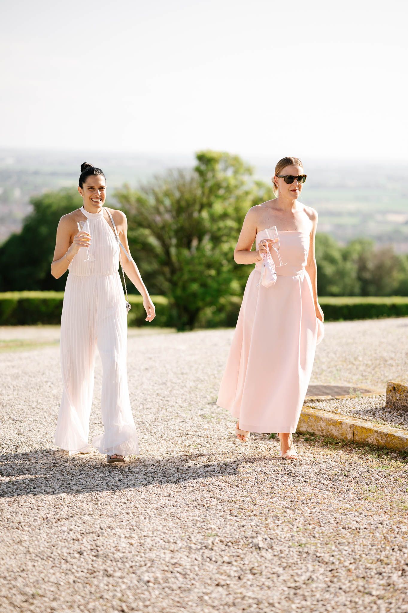 Two attendants in blush pink dresses walk along a gravel path with champagne glasses.