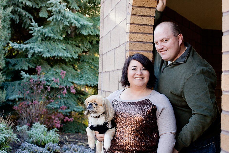 Couple poses with their small Pekingese dog in front of a landscaped garden.