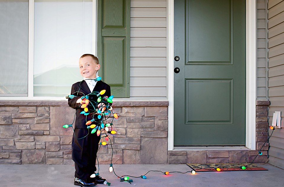 Small child wrapped in colorful Christmas lights standing on a front porch.