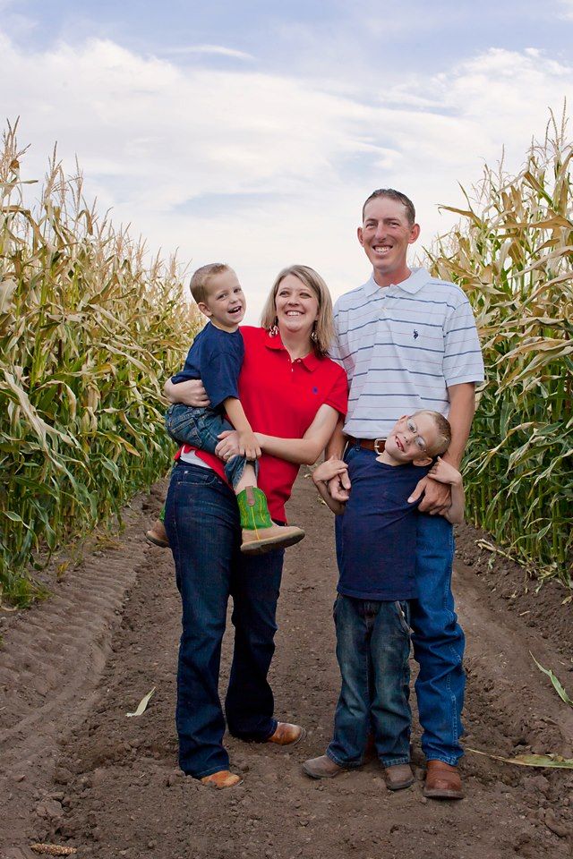 Family of four posing together between tall corn stalks on a farm.