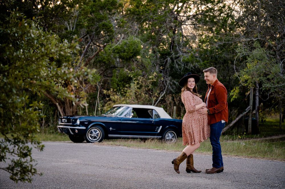 A couple poses together near a classic black car on a dirt path surrounded by autumn trees.