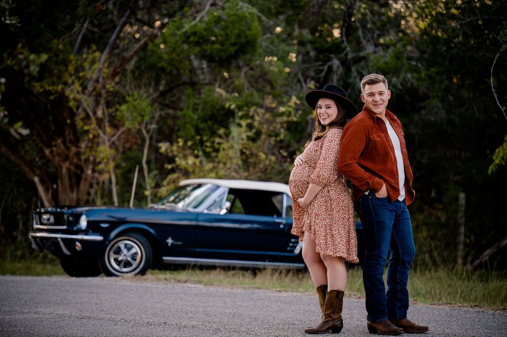 Maternity portrait with vintage blue Mustang car in wooded setting at sunset.
