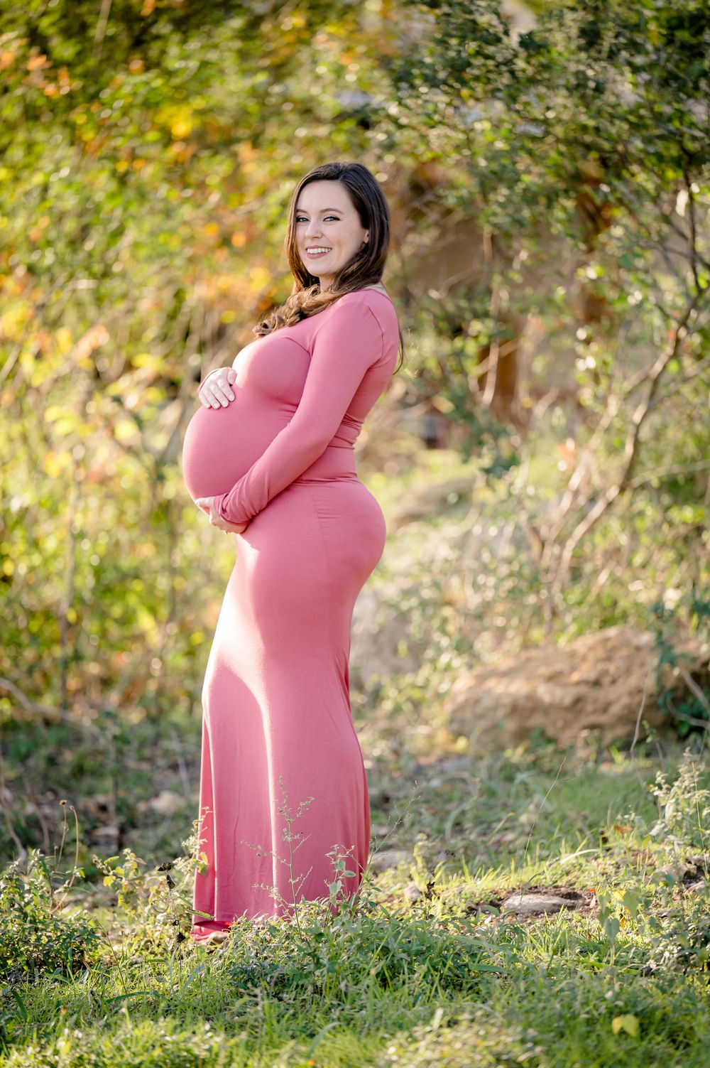 An expectant mother in a pink dress poses in a natural outdoor setting during golden hour.