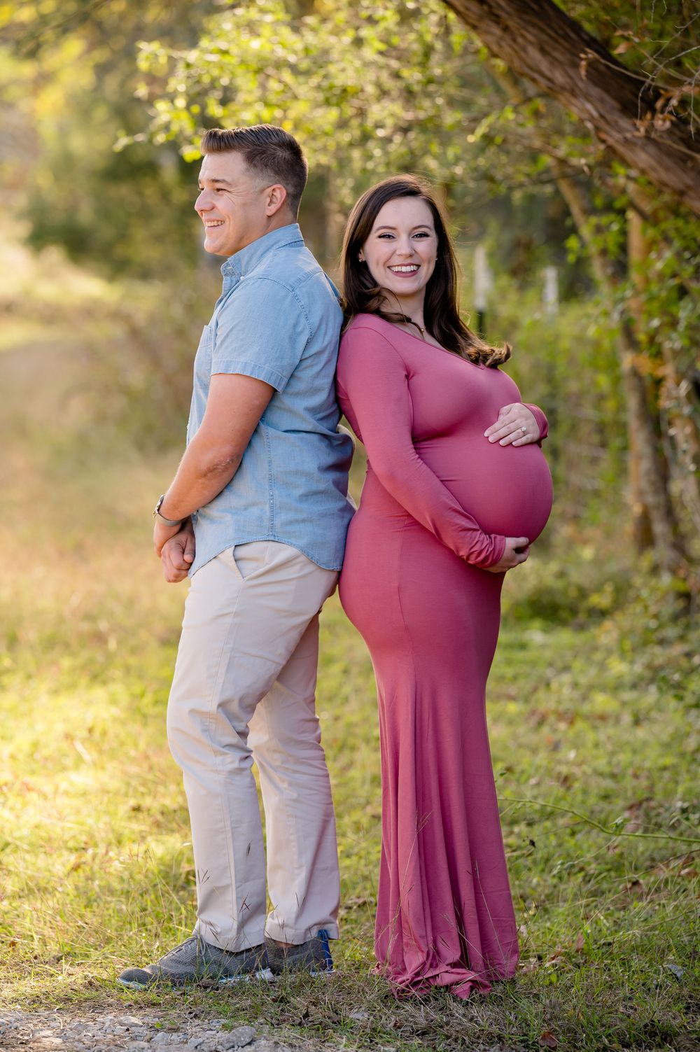 A couple poses back-to-back outdoors during a maternity photo session in warm evening light.
