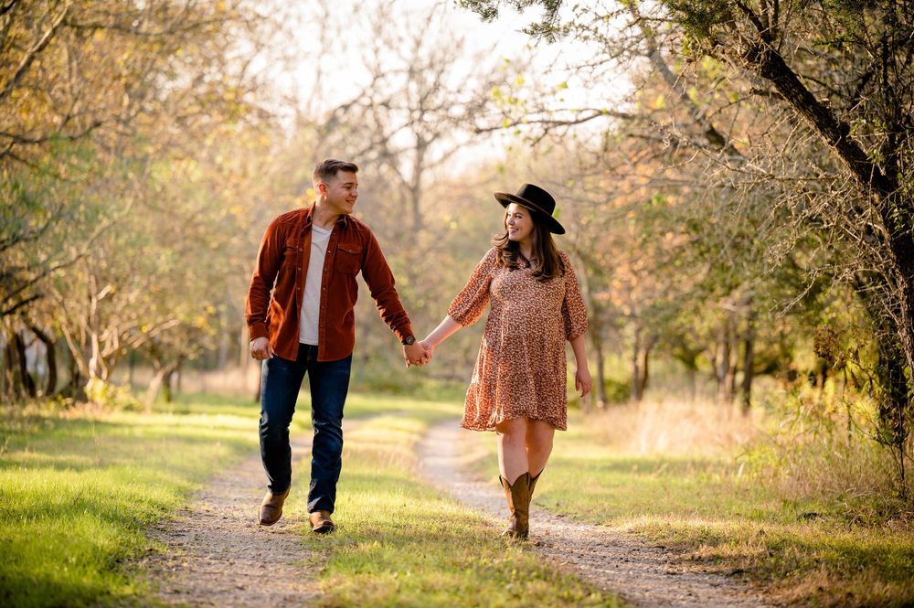 Couple holds hands while walking together on wooded trail during sunset maternity session.