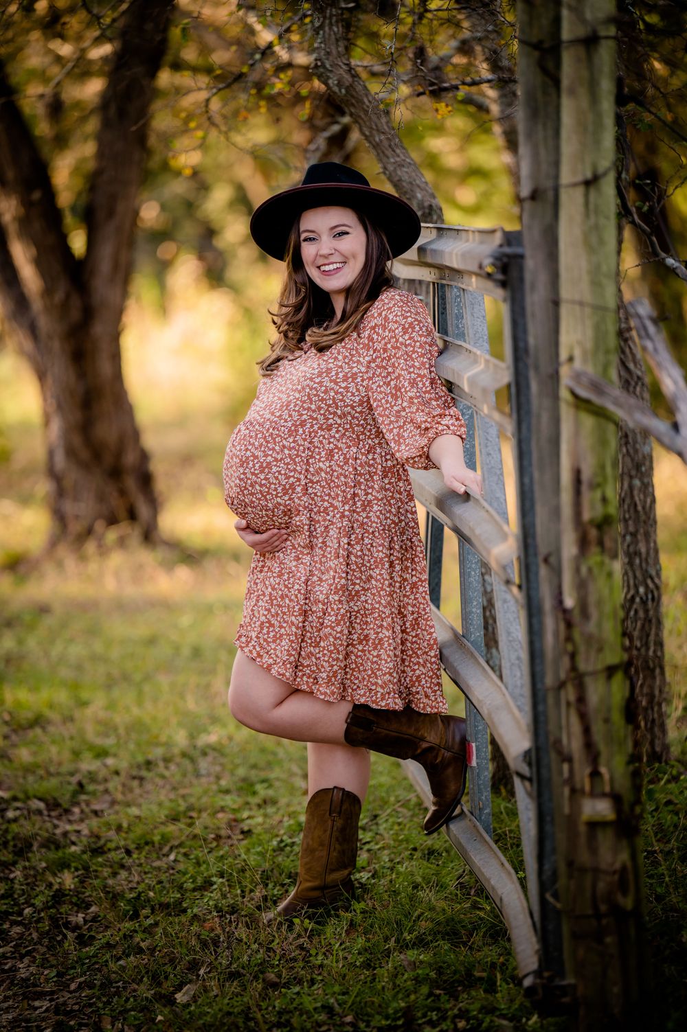 Pregnant woman in floral dress and cowboy boots leans against rustic fence in autumn setting.