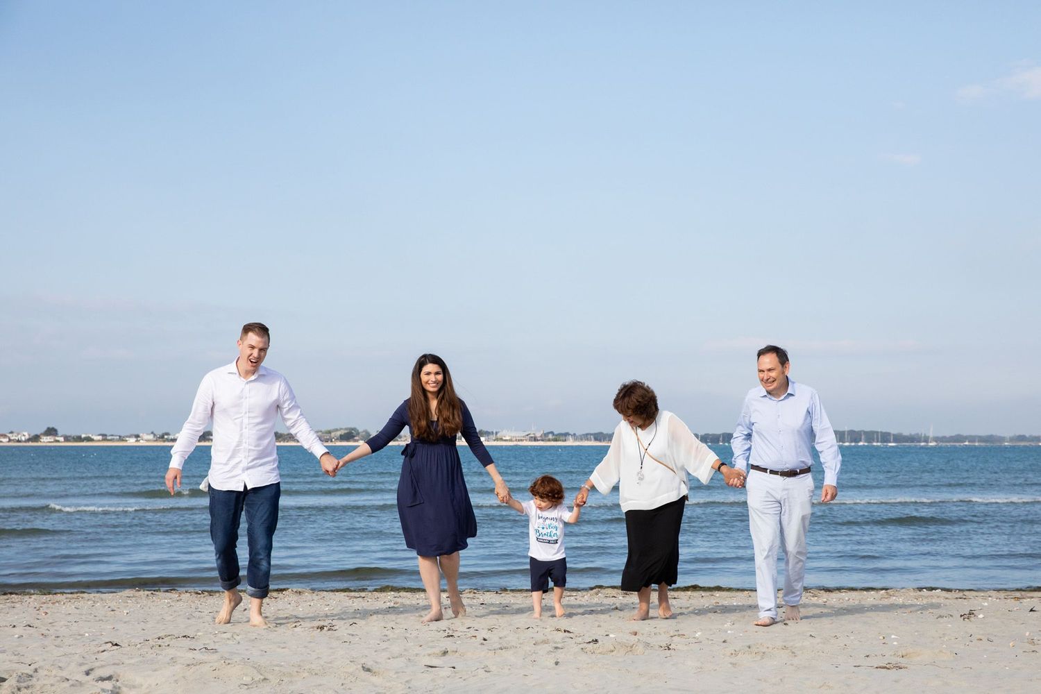 A family walks hand-in-hand along a beach with ocean and blue sky in the background.