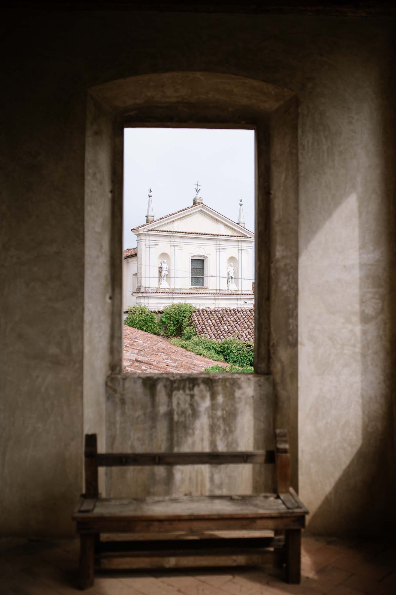 View of a white church facade through a rustic stone window frame with wooden bench.