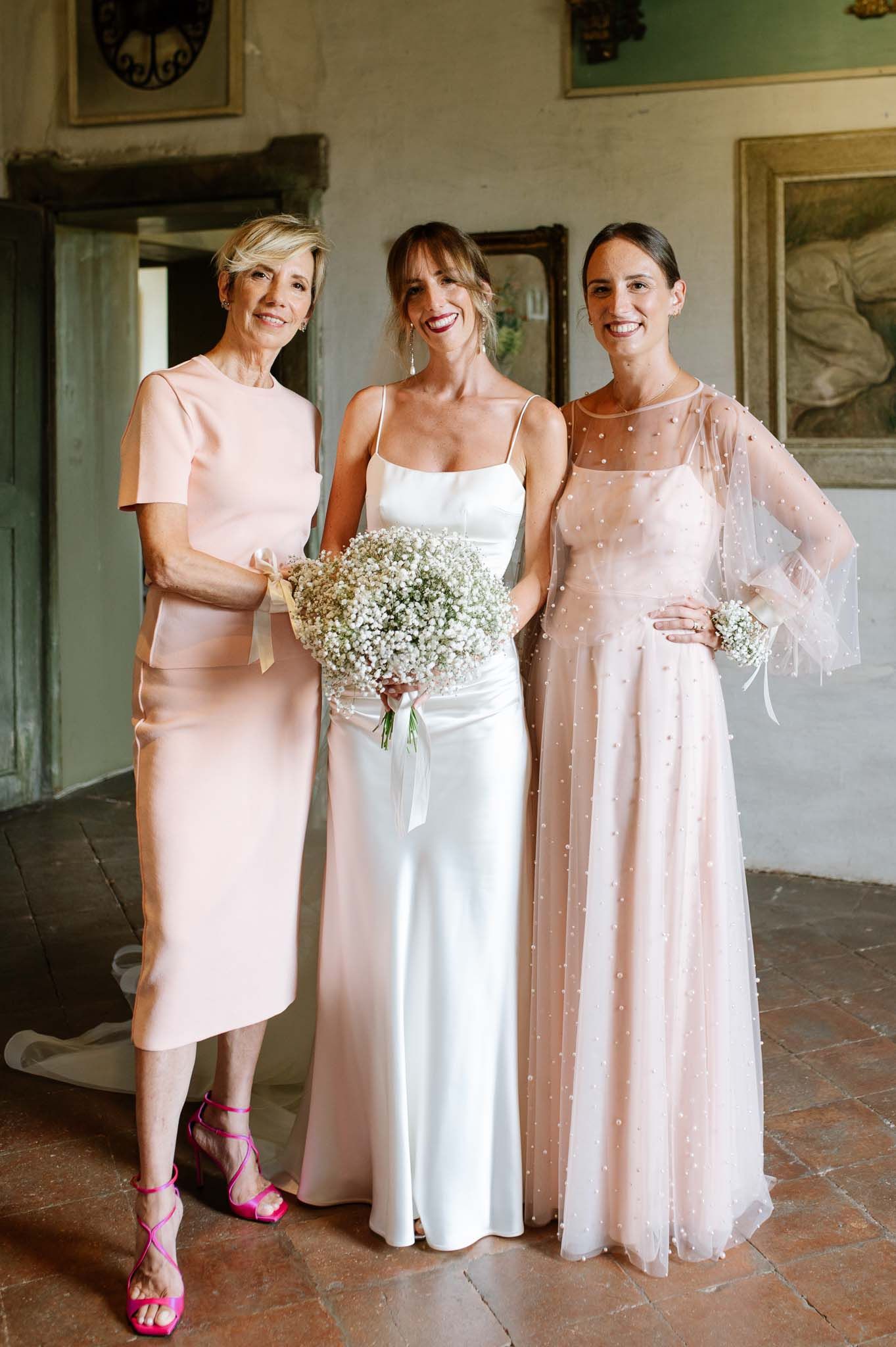 Three women in pastel pink dresses pose together holding baby's breath bouquets.