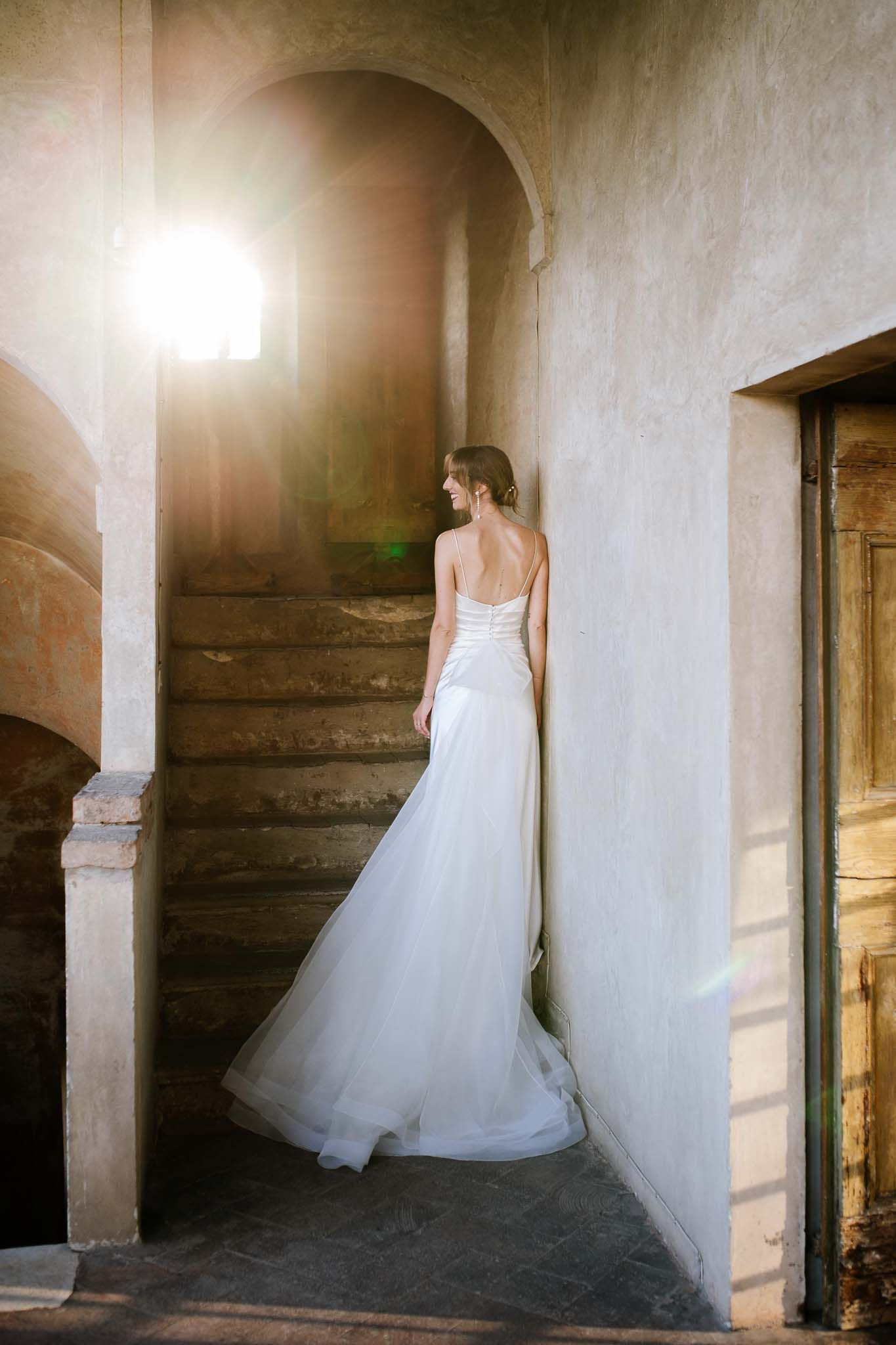 Bride in flowing white dress stands elegantly on stone stairs with sunlight streaming through arched windows.