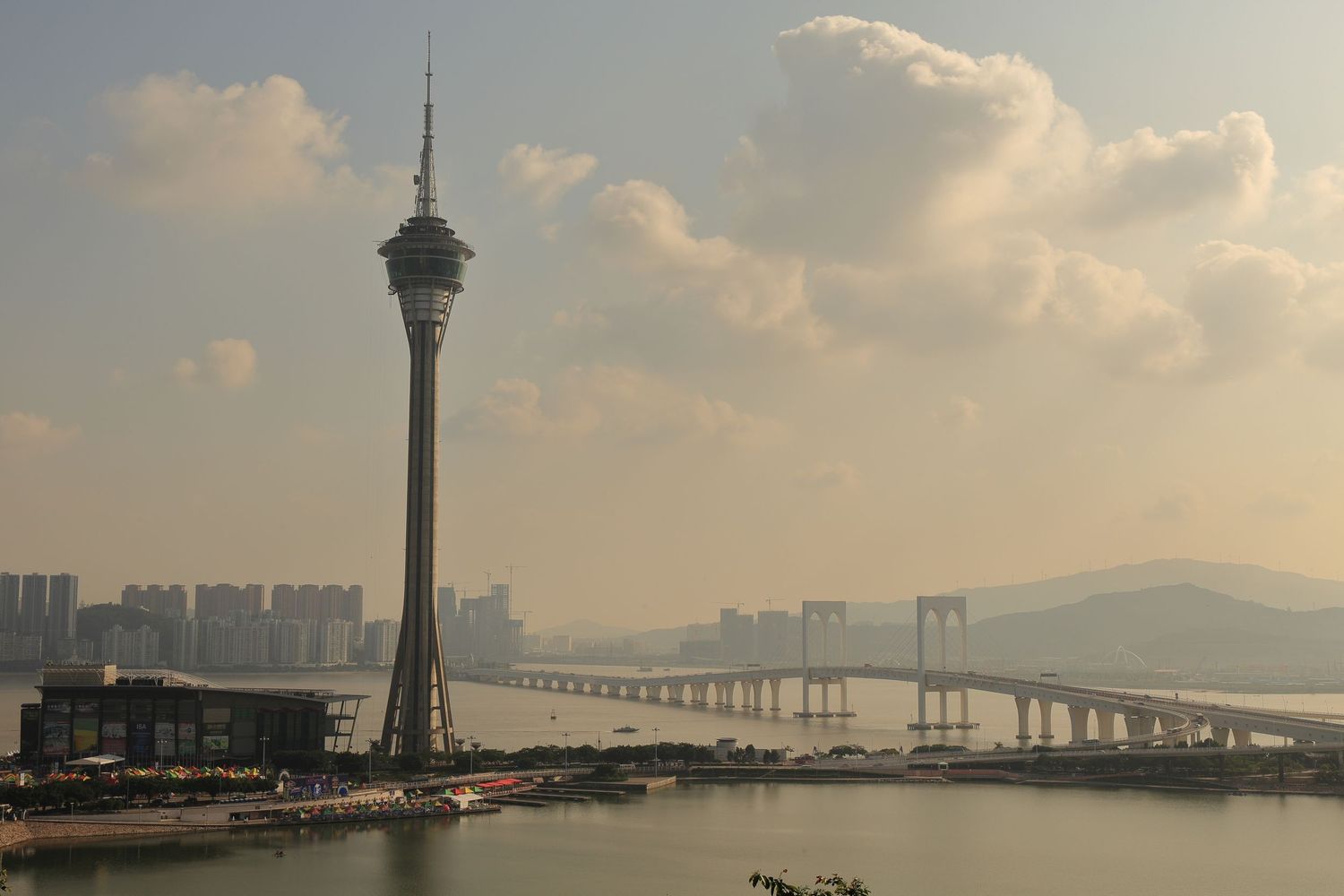 Macau Tower and bridge silhouetted against hazy sunset sky over water.