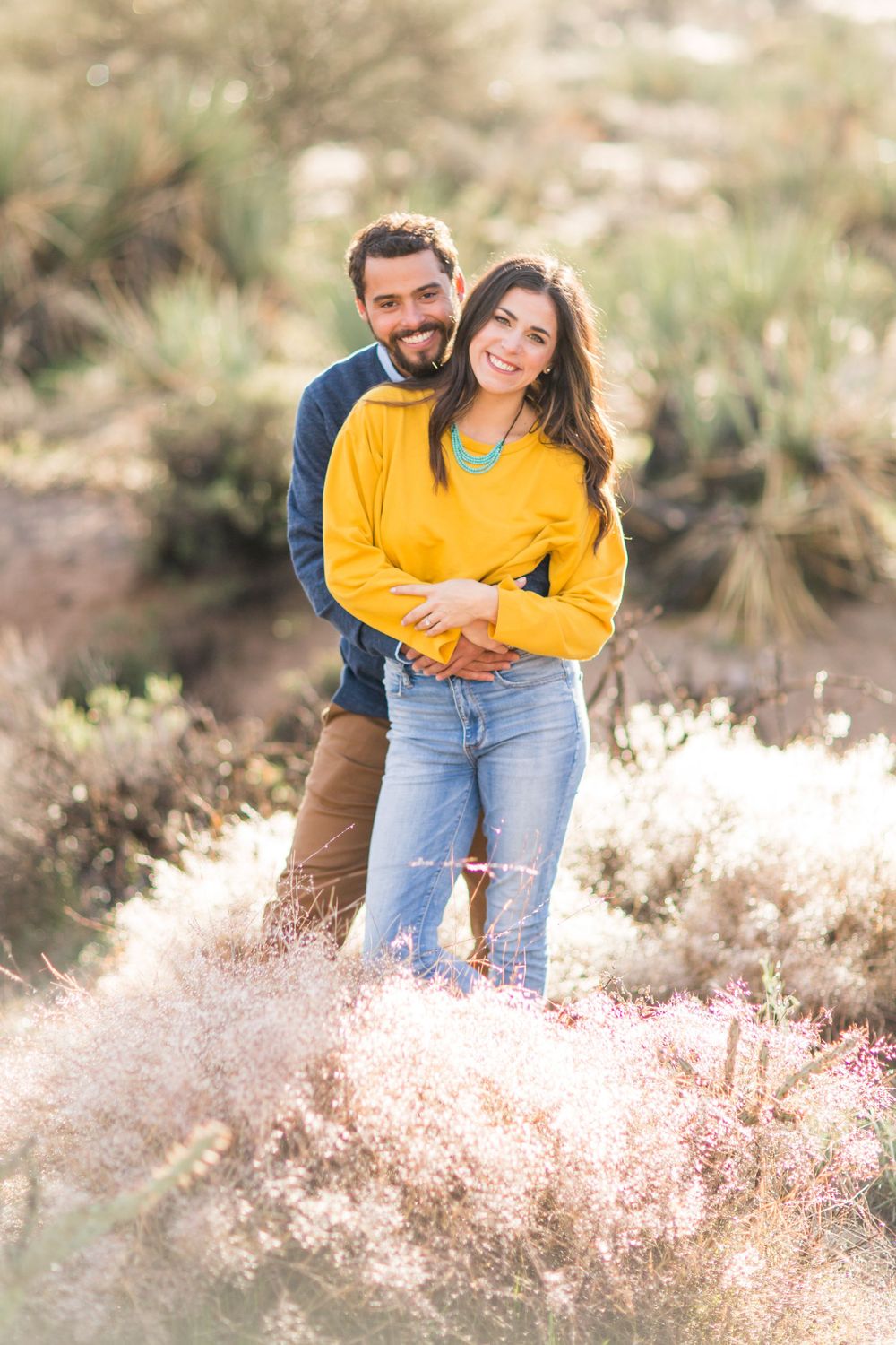 Couple in yellow and navy sweaters embracing in a desert landscape with flowering plants.