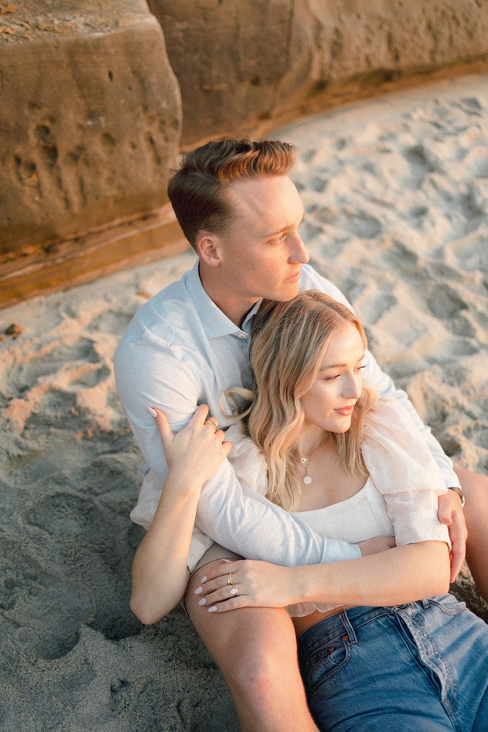 Couple relaxes together on sandy beach against weathered cliff wall.