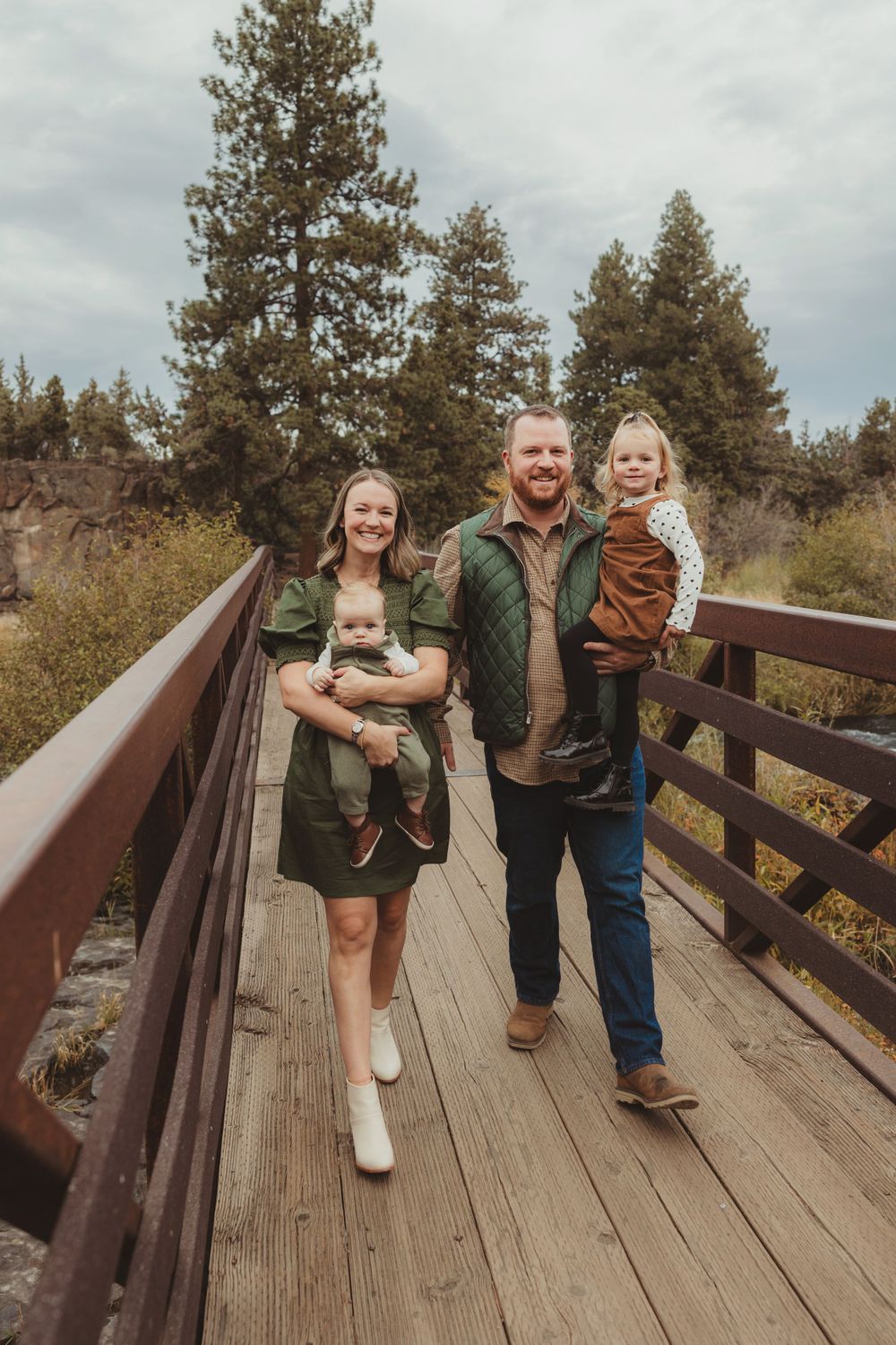 A family poses on a wooden bridge with towering pine trees and cloudy skies in the background.