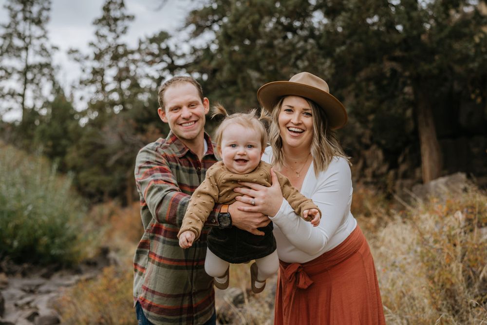 A happy family of three poses together on an autumn trail surrounded by trees.