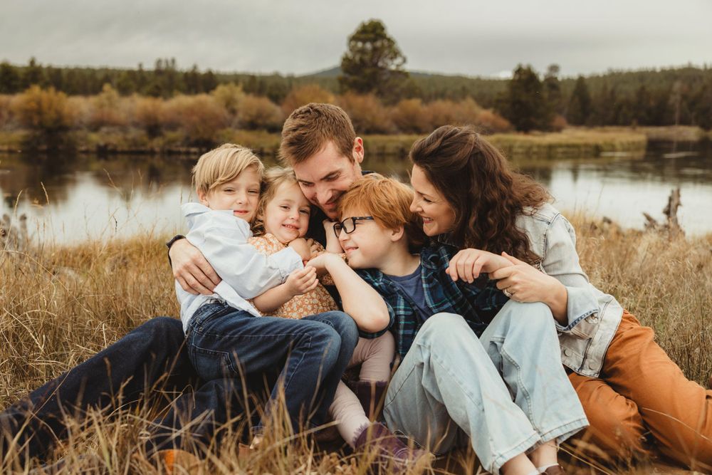 Family cuddles together in tall grass by a serene lake during golden hour.