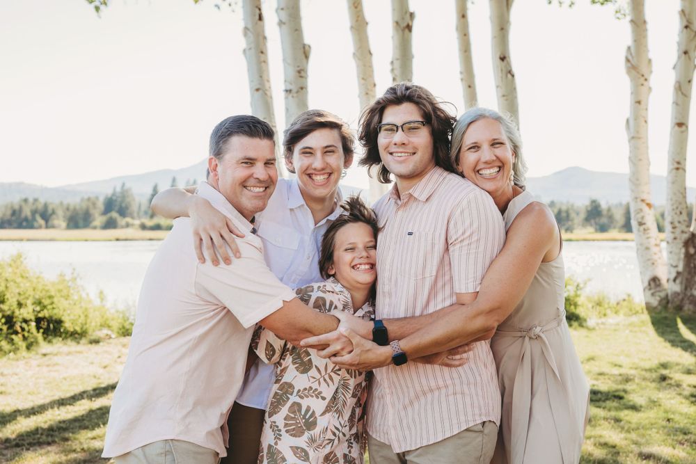 A family embraces while standing in front of white birch trees and mountains during summer.