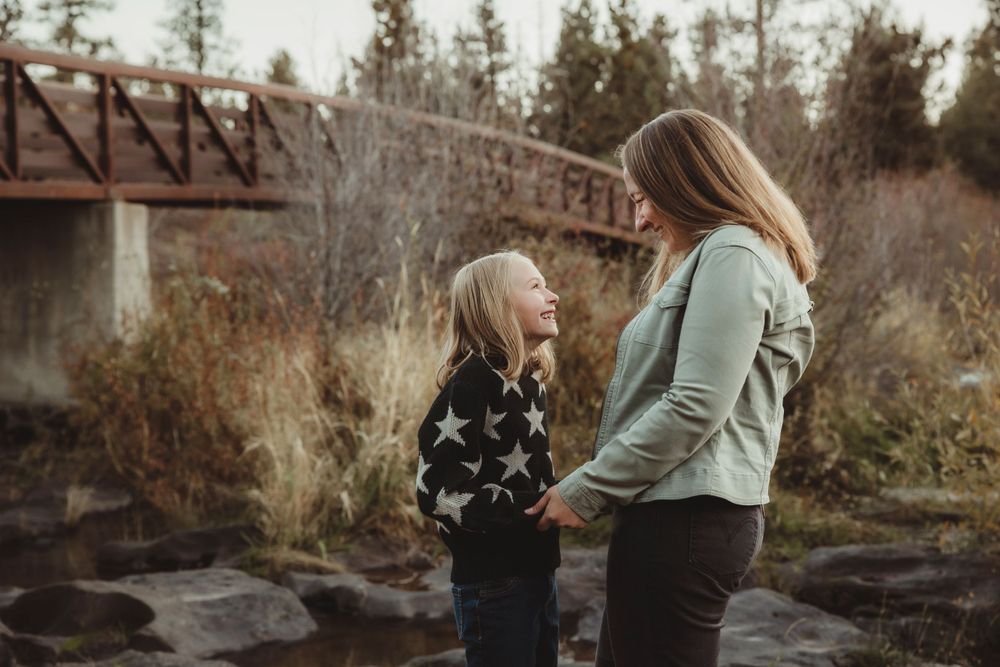 A mom and daughter share a candid moment near a bridge in muted autumn tones.
