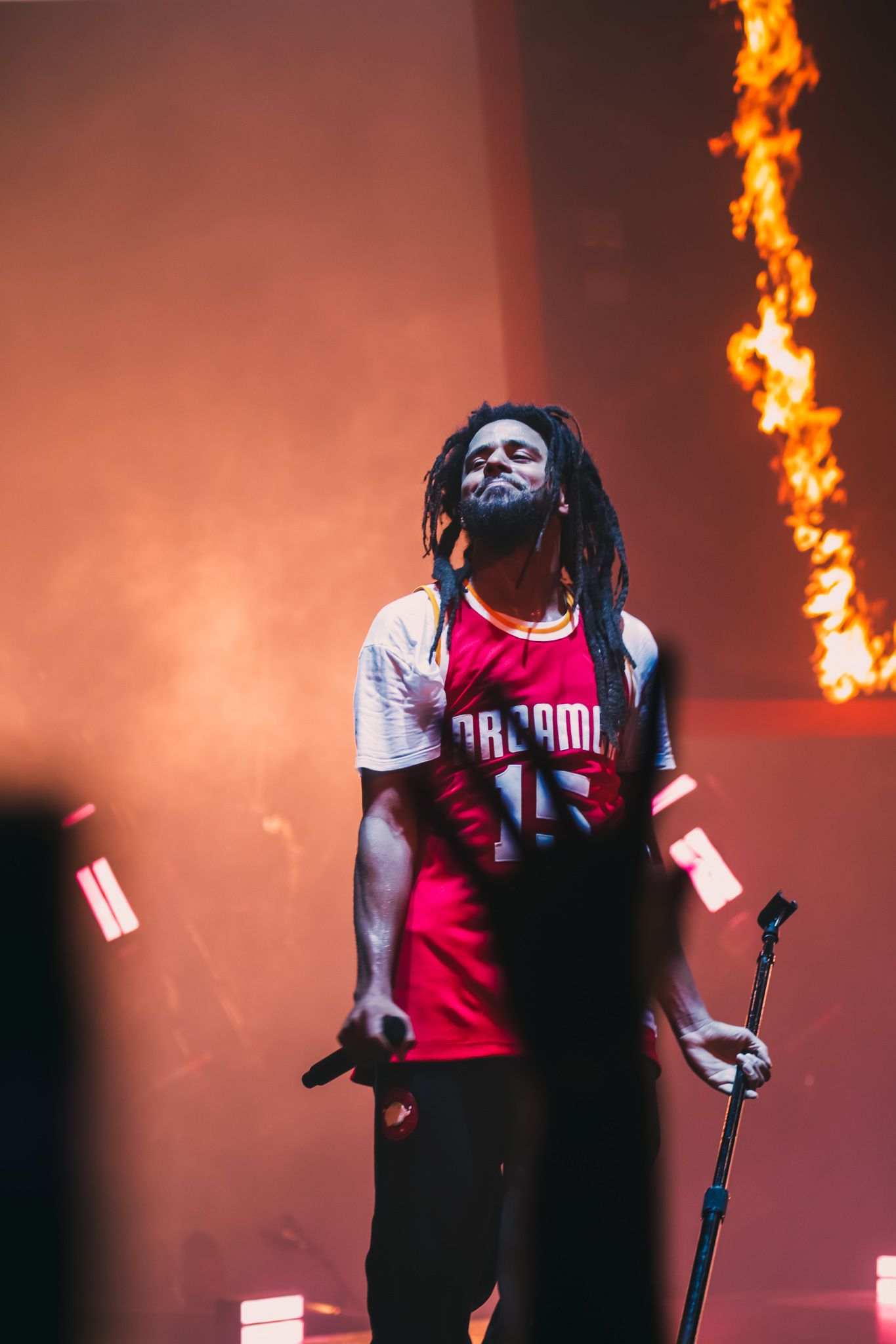 Artist performs on stage wearing a red jersey with dramatic fire effects in the background.