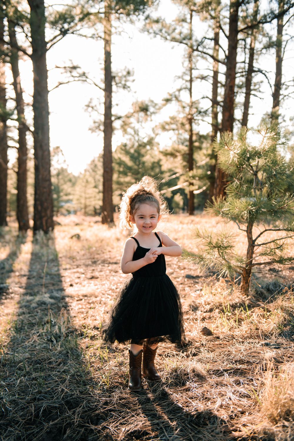 Young child in black dress twirls among pine trees at sunset.