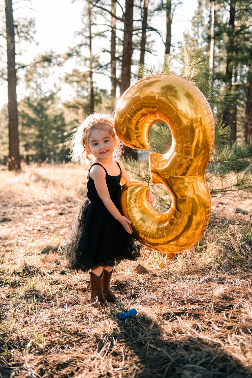 Child plays with large gold number 3 balloon in pine forest setting.