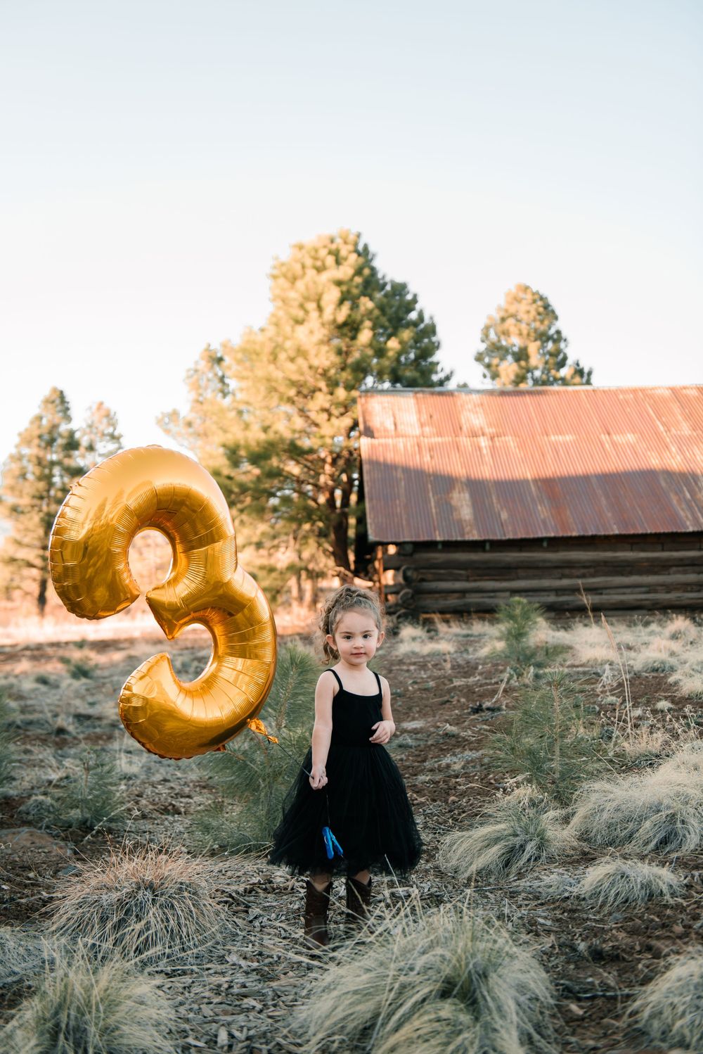 Child in black dress dances with gold number balloon in outdoor setting.