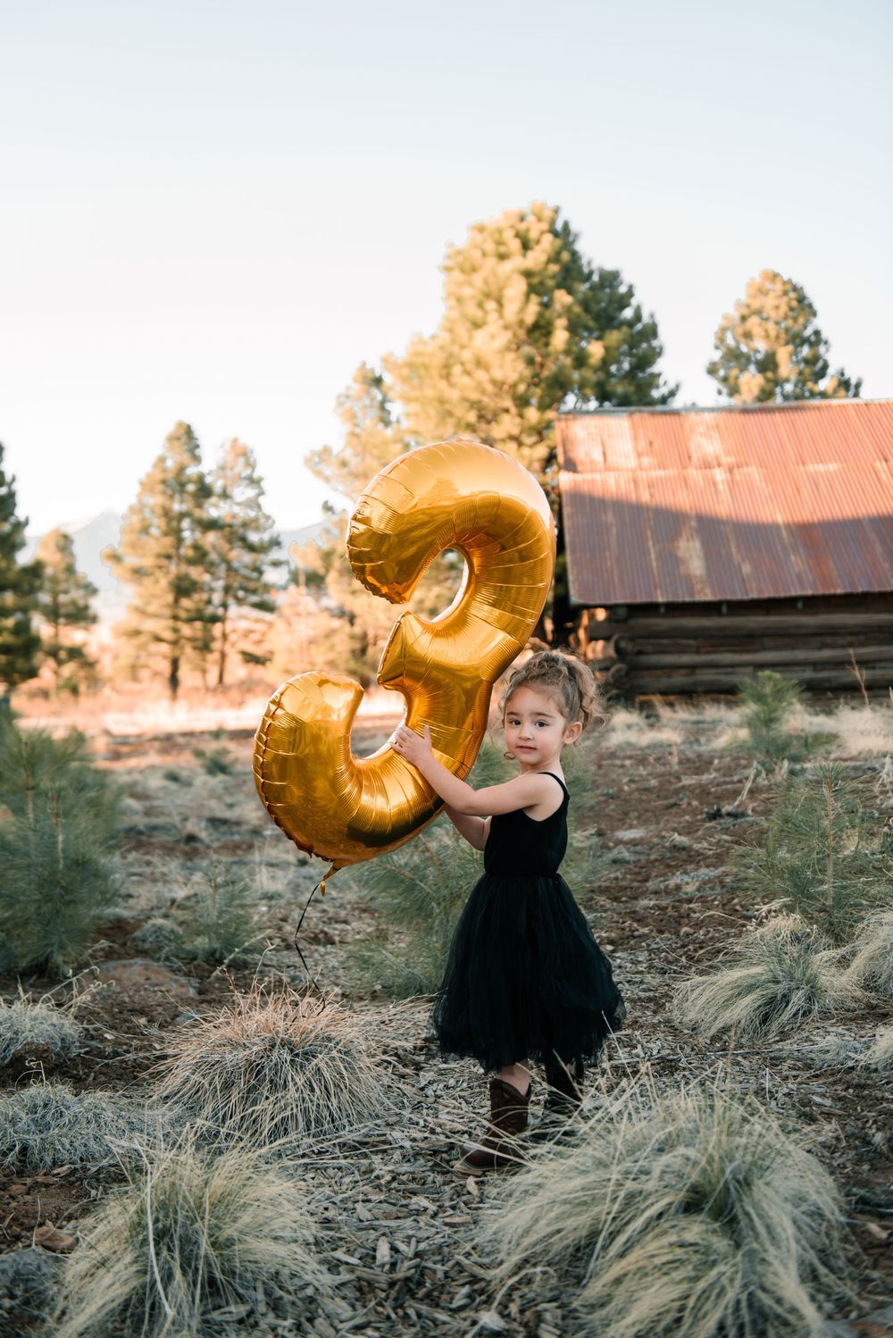Child holding gold number 3 balloon poses near rustic building at sunset.