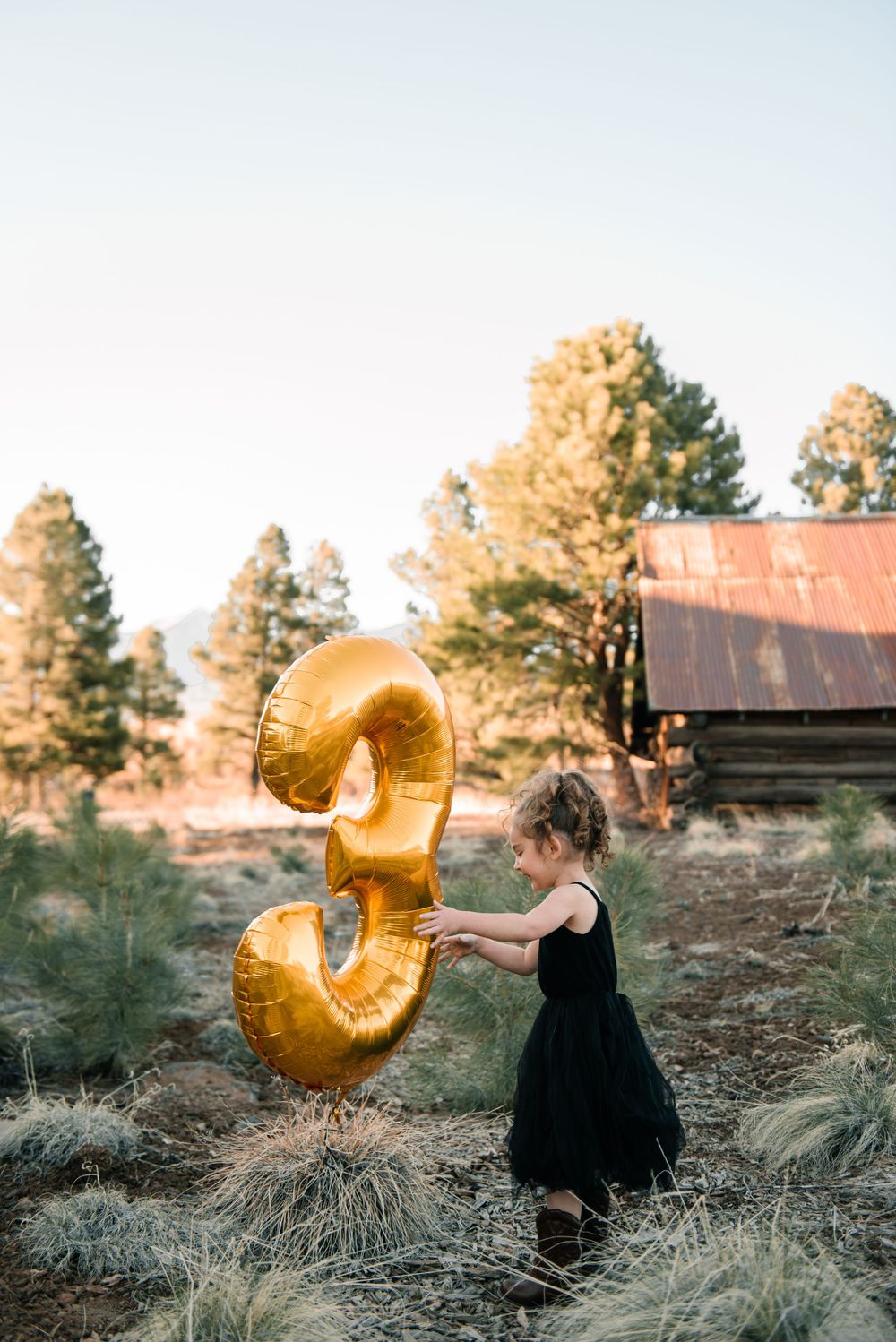 Child twirls with golden number 3 balloon against sunset forest backdrop.