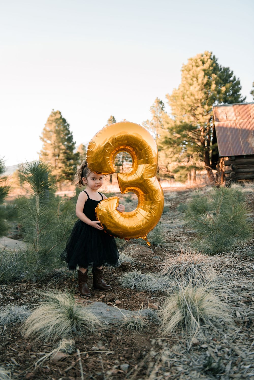 Someone wearing black holds a shiny gold number 9 balloon near a rustic wooden structure.