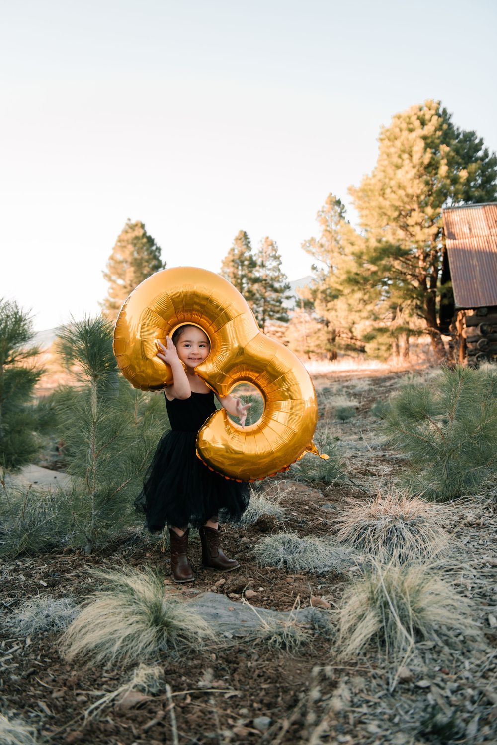 A figure in black holds a large golden number 9 balloon against a rustic outdoor backdrop at sunset.