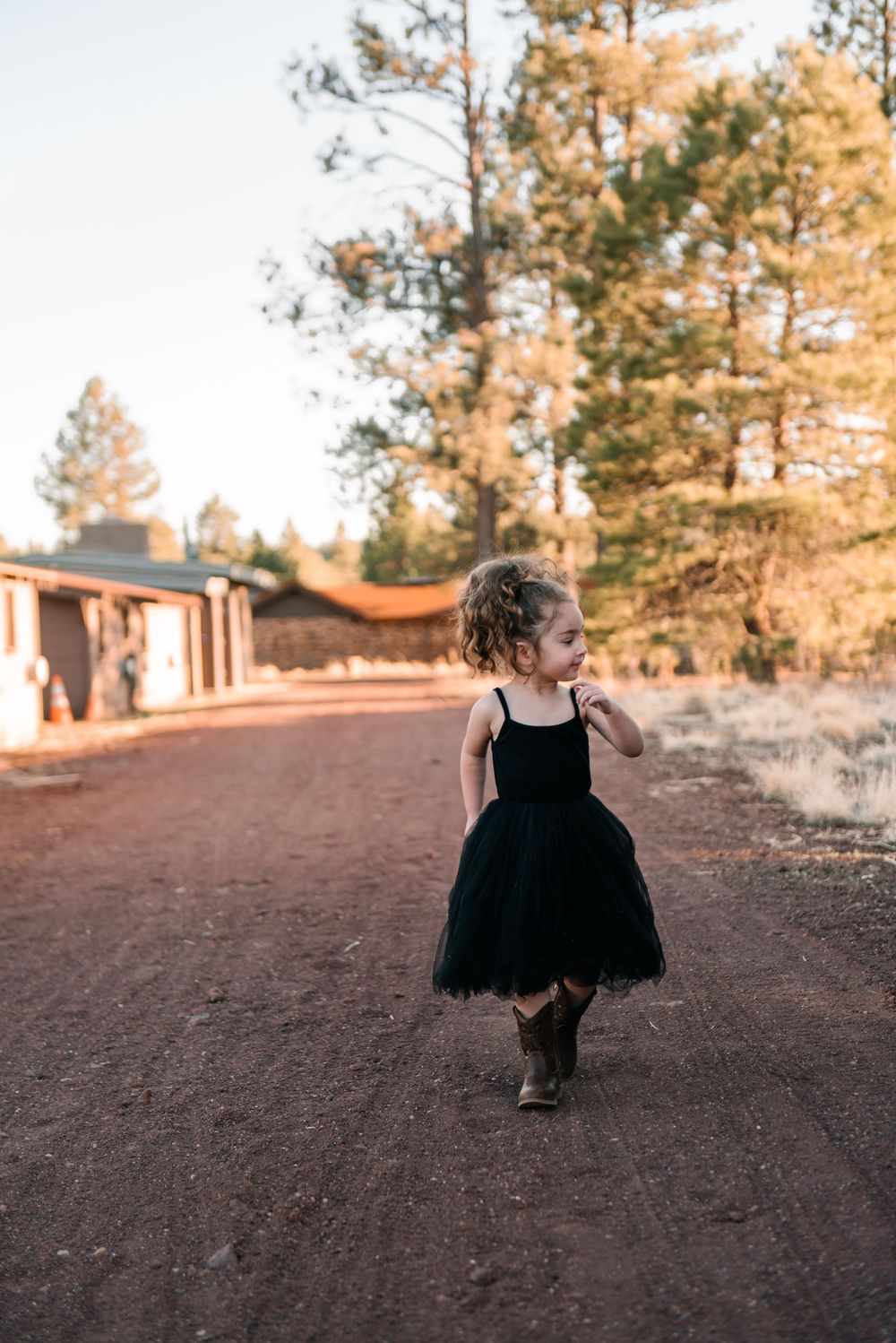 A small figure in black twirls on a dirt path with pine trees in the background at sunset.