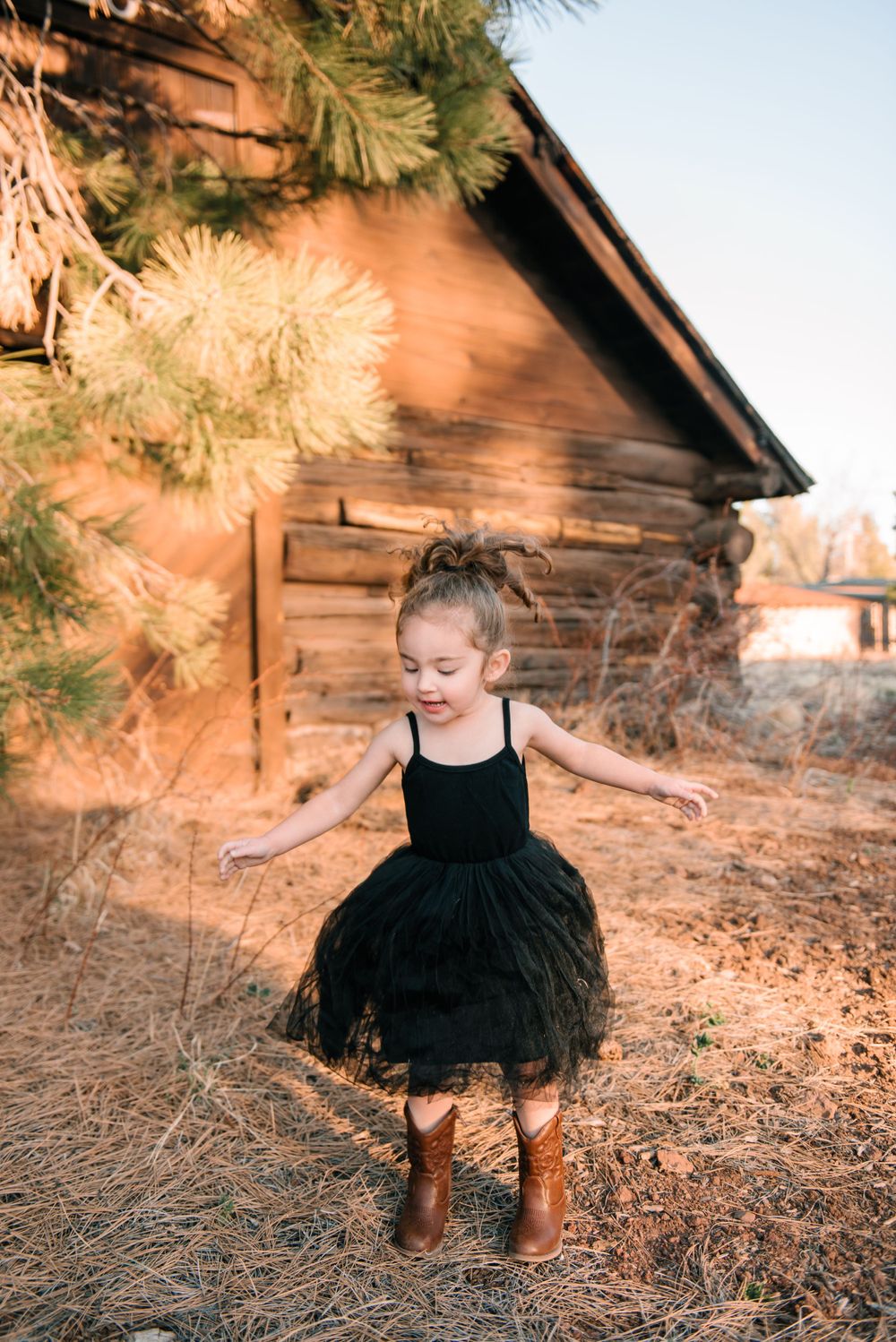 A young figure in a black tulle dress dances near a rustic cabin during golden hour.