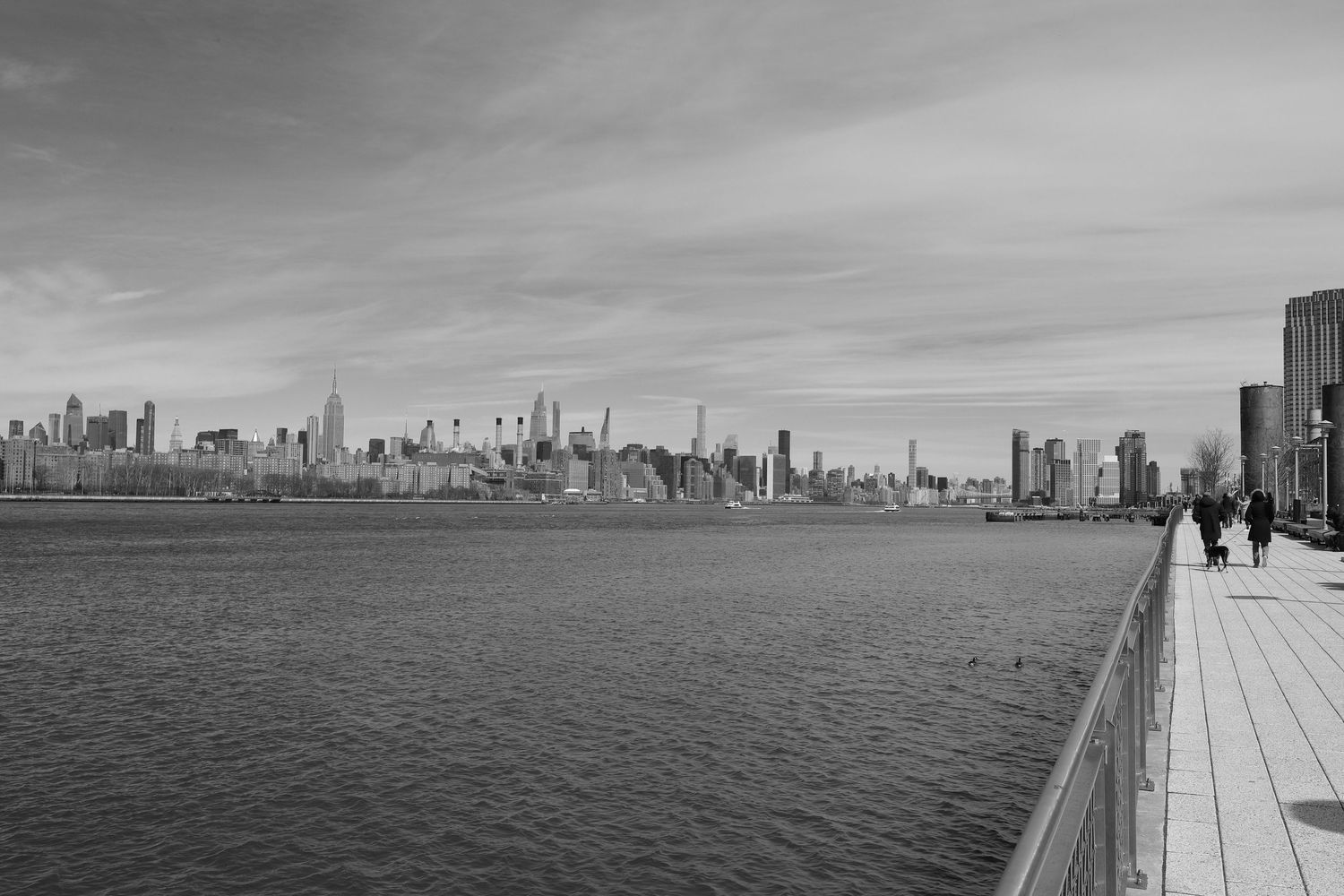 NYC skyline viewed from the waterfront with pedestrians walking along the river's edge.
