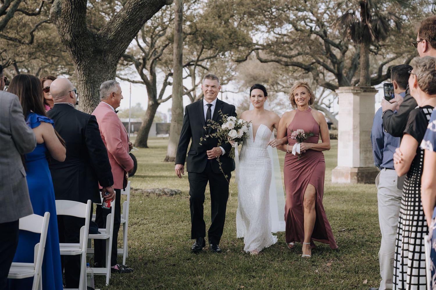 Wedding party walks down outdoor aisle beneath old trees with guests seated on white chairs.